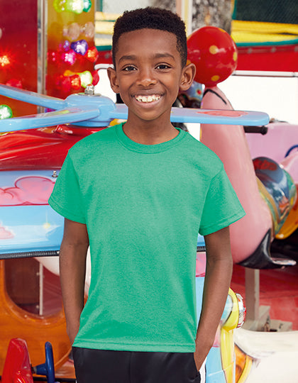 En ung dreng iført en grøn T-shirt fra Fruit of the Loom smiler, mens han står foran en farverig forlystelse med blinkende lys på en forlystelsespark.