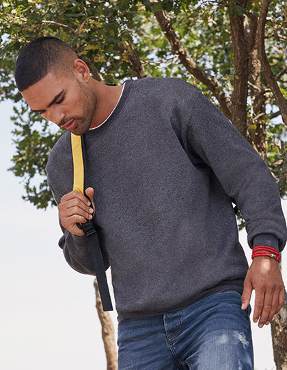 En mand iført en mørkegrå Fruit of the Loom-trøje og blå jeans går udenfor, bærer på en taske med gult rem, omgivet af træer i baggrunden.