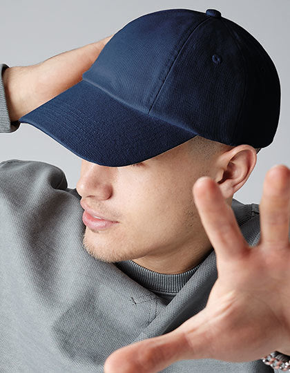 En person med en mørkeblå kasket laver en håndbevægelse nær ansigtet, iført en grå sweatshirt fra Fruit of the Loom, i en neutral indendørs setting.