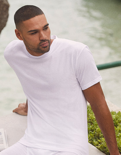 Mand sidder ved en flod, klædt i en hvid T-shirt fra Fruit of the Loom, med én hånd hvilende på en stenbænk nær grønne planter og et vandmiljø i baggrunden.